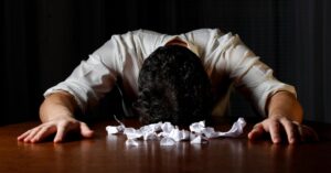 person with their head on a desk surrounded by paper balls
