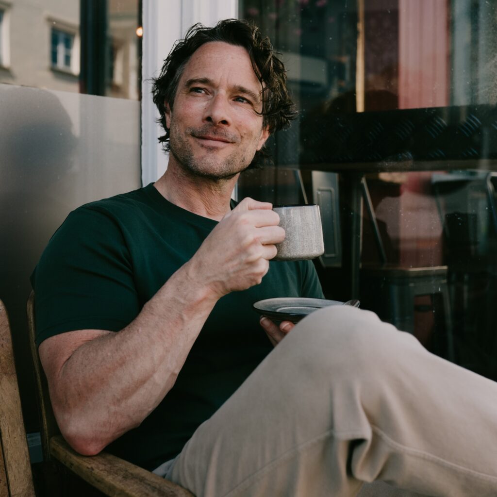 Man with dark hair in a black shirt sits by a window, holding a gray mug and smiling slightly at the camera.