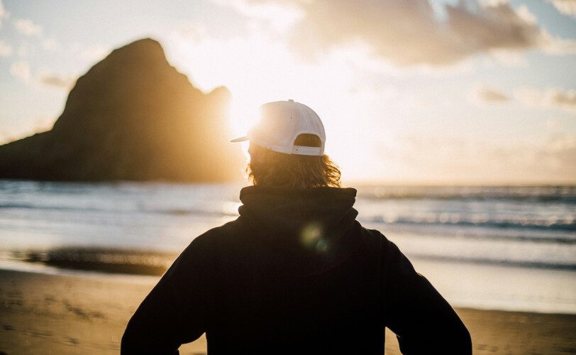 Person in a hoodie and white cap stands on a sandy beach at sunset, facing the sea toward a distant rocky island.