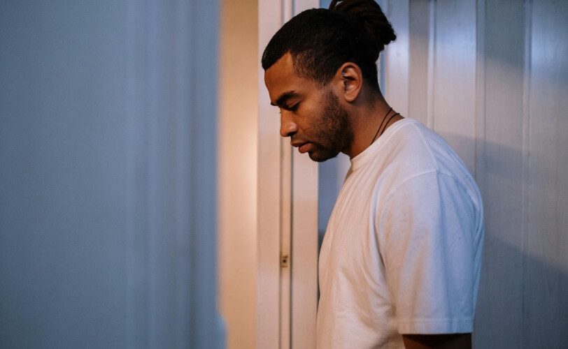 Side-profile of a man with a bun wearing a white t-shirt, leaning against a blue wall in a hallway, looking down pensively.